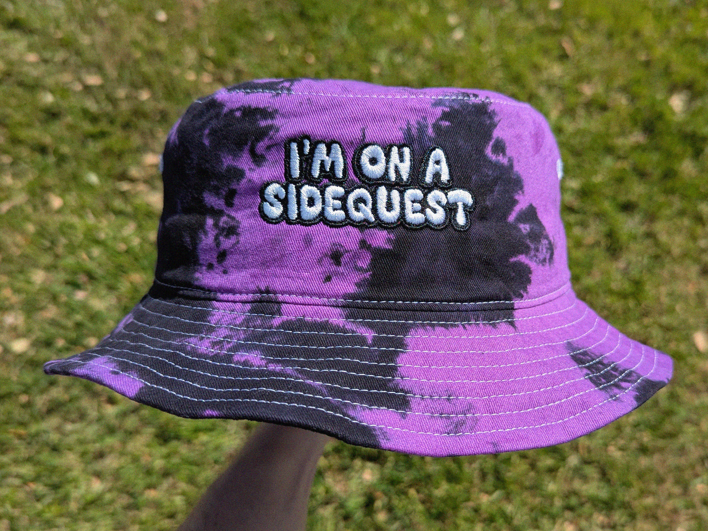 A tie-dye bucket hat with 'I'm on a sidequest' embroidered on it, predominantly purple with black patterns.