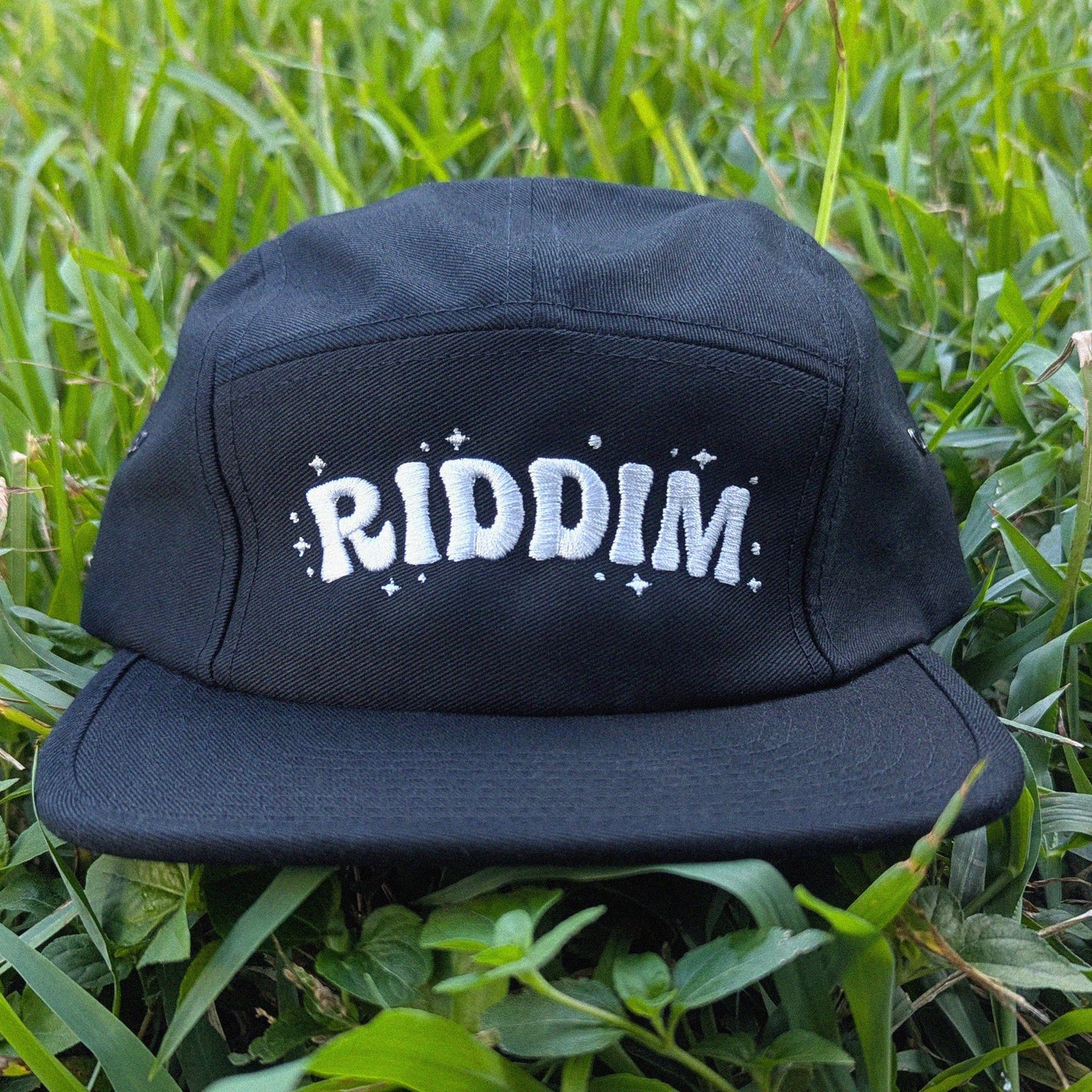 A black flat-brim cap with 'RIDDIM' embroidered in white across the front, resting on green grass.
