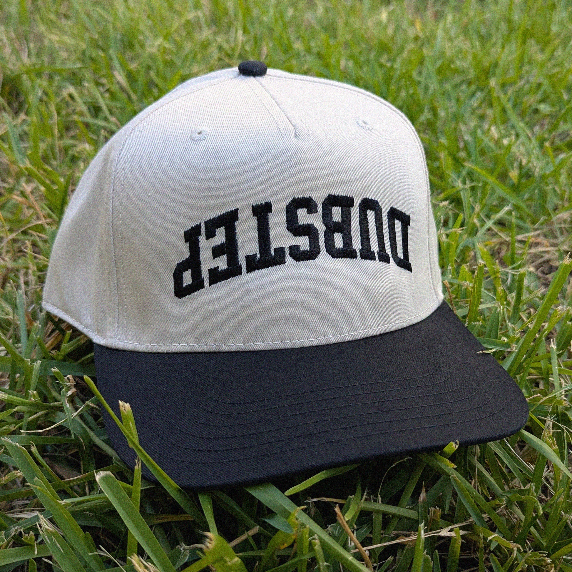 A two-tone baseball cap with an off-white crown and a black flat brim, featuring the word 'DUBSTEP' embroidered in black across the front. The cap is photographed resting on green grass.