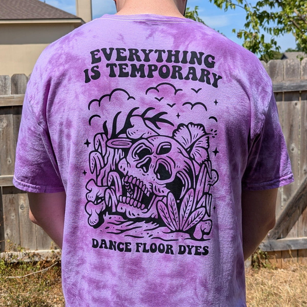 A person viewed from behind wearing a purple tie-dye t-shirt with a large black graphic of a skull and flowers and the text 'EVERYTHING IS TEMPORARY' and 'DANCE FLOOR DYES'.