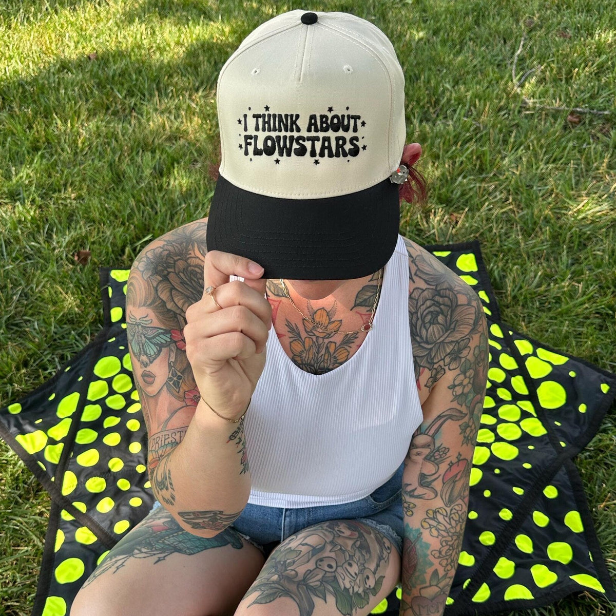 A tattooed person sitting on green grass outdoors, wearing a black and off-white baseball cap with the text 'I THINK ABOUT FLOWSTARS' and a white tank top, with a black and neon yellow patterned blanket underneath.