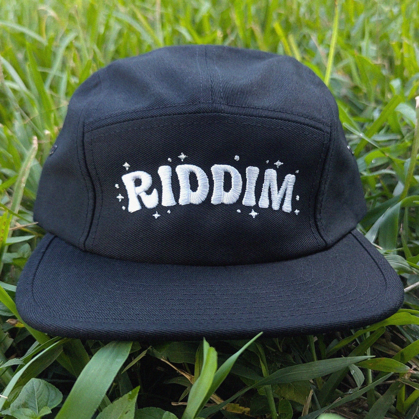 A black 5-panel camp cap with 'RIDDIM' embroidered in white on the front, resting on green grass.