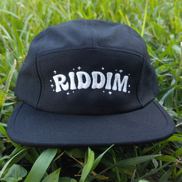 A black 5-panel camp cap with 'RIDDIM' embroidered in white on the front, resting on green grass.