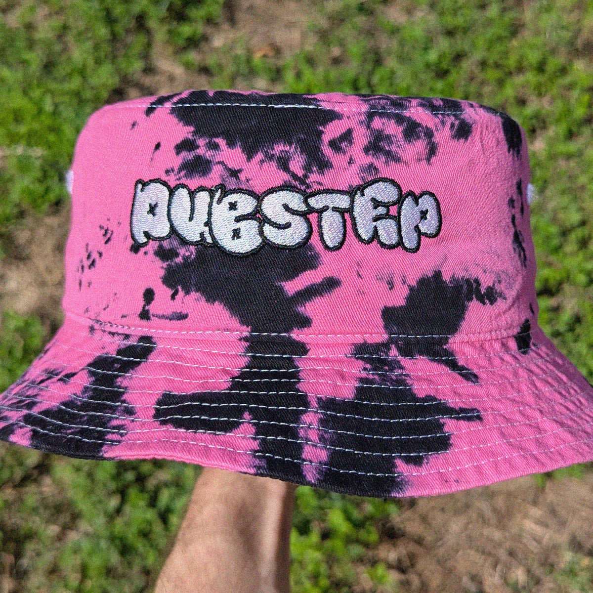 A pink and black tie-dye bucket hat with 'DUBSTEP' embroidered in white and black across the front, held up by a hand against a blurry green grass background.