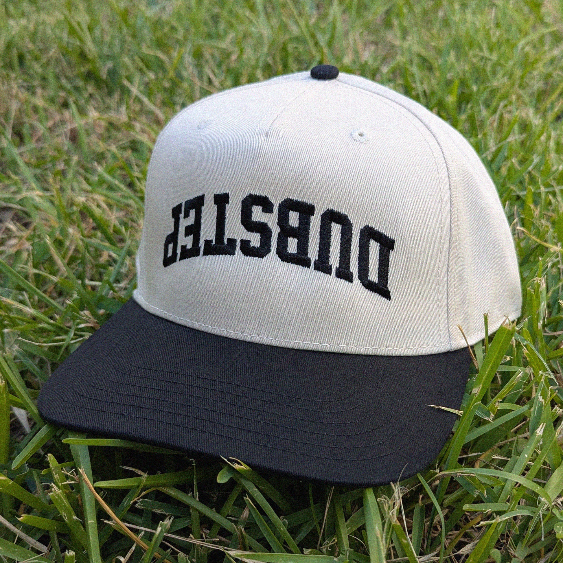 A two-tone baseball cap with an off-white crown and a black flat brim, resting on green grass. The front of the cap features the word 'DUBSTEP' embroidered in black arched block letters.