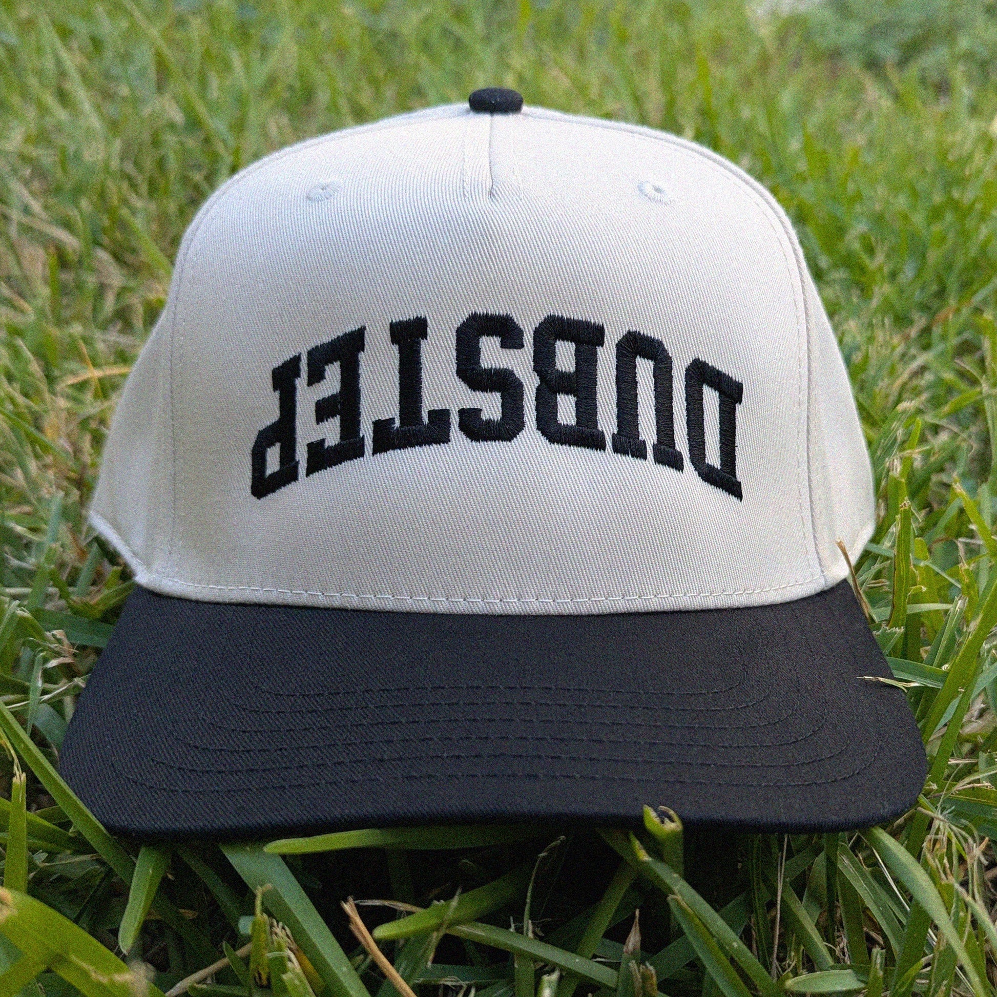 A black and tan snapback cap with 'DUBSTEP' embroidered in black arched letters on the front, resting on green grass.