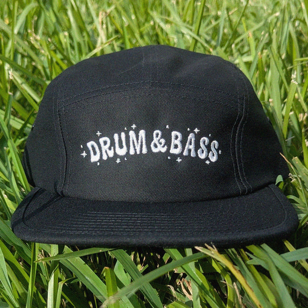 A black five-panel cap with 'DRUM & BASS' embroidered in white across the front, resting on green grass.