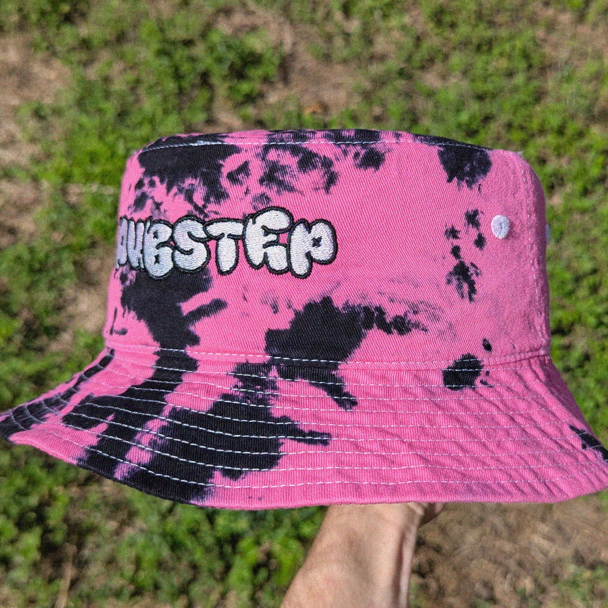A pink and black tie-dye bucket hat with the word 'DUBSTEP' embroidered on the front, held up by a hand against a background of green grass.