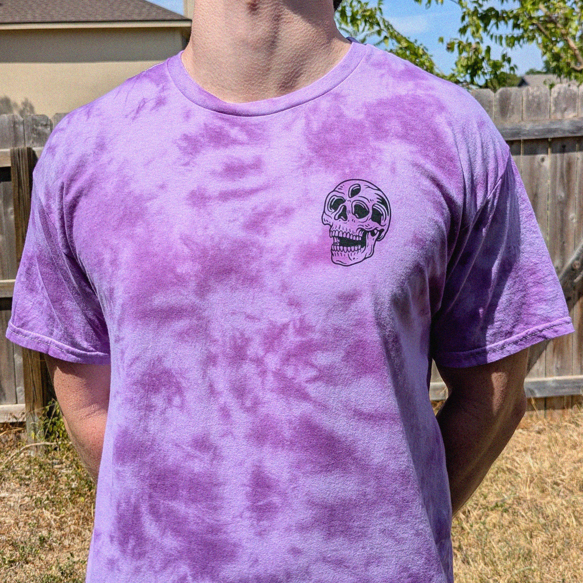 A person wearing a purple tie-dye t-shirt with a small black skull graphic on the left chest, photographed outdoors in front of a wooden fence.