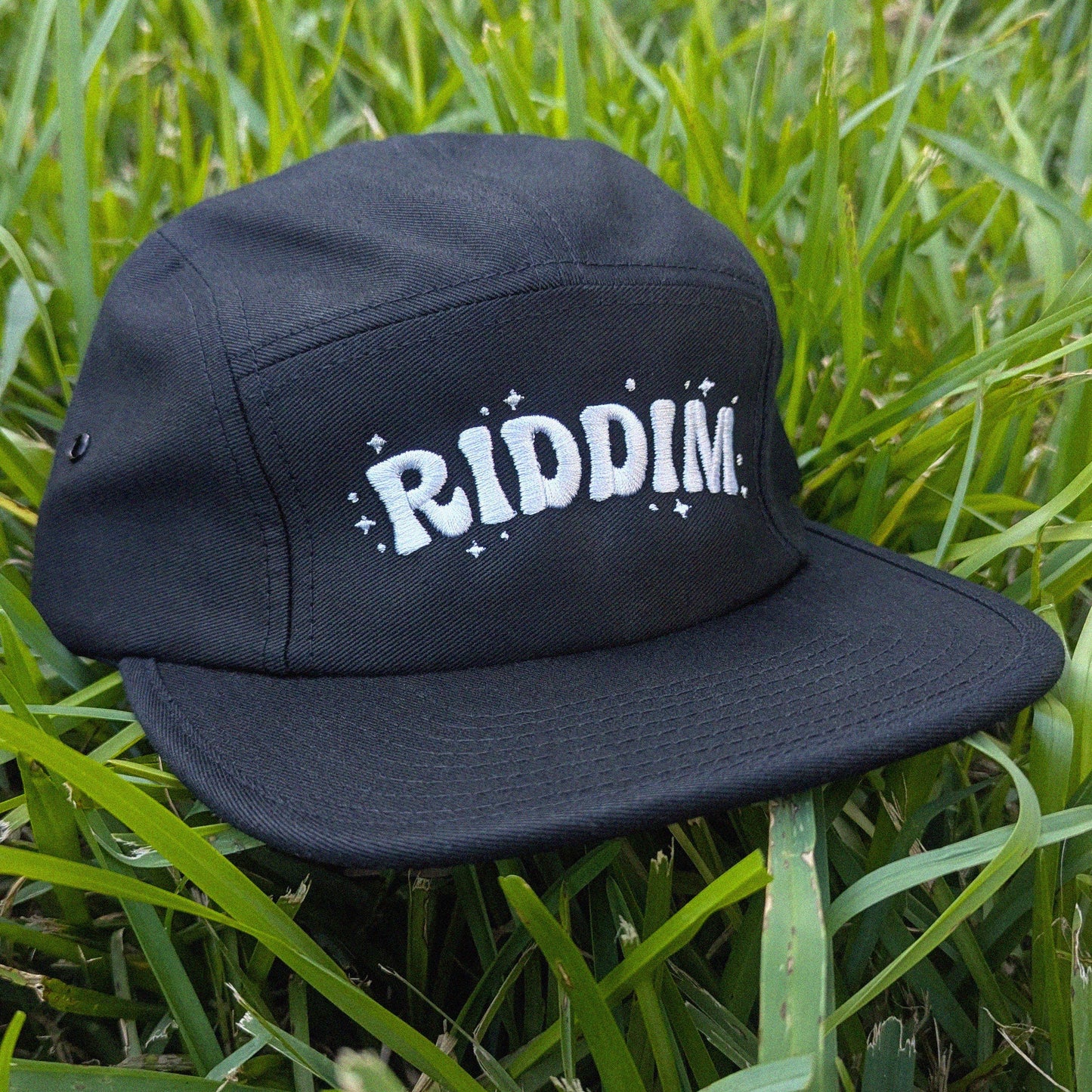 A black five-panel cap with 'RIDDIM.' in white retro-style lettering and small white star accents, resting on green grass.