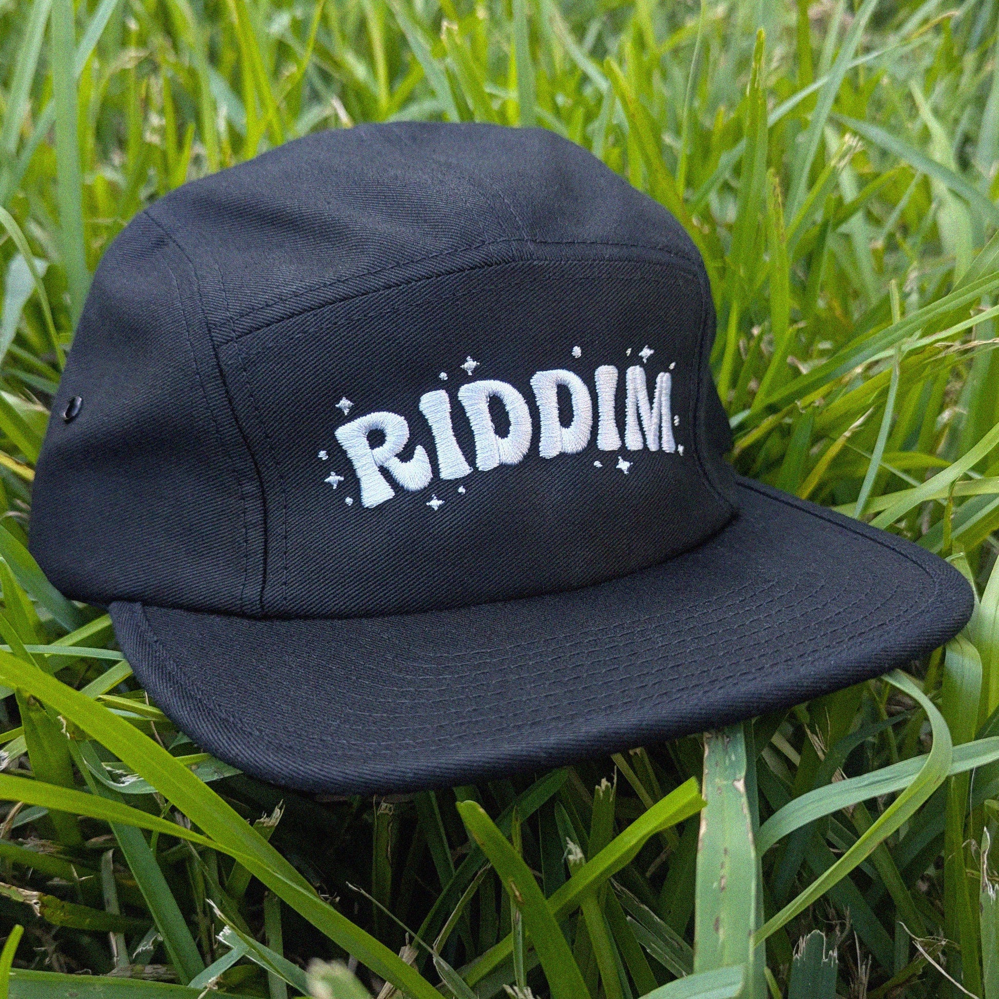 A black five-panel cap with 'RIDDIM.' in white retro-style lettering and small white star accents, resting on green grass.