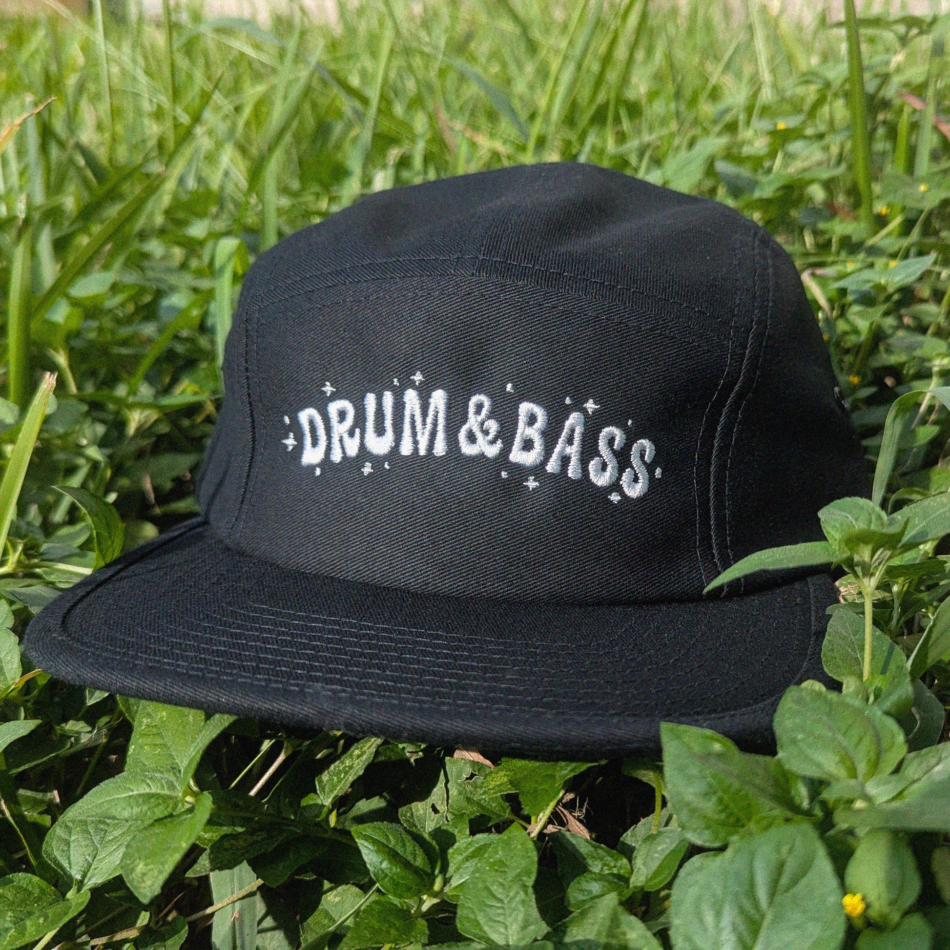 A black flat-brim cap with 'DRUM & BASS' embroidered in white on the front, resting on green grass and foliage outdoors.