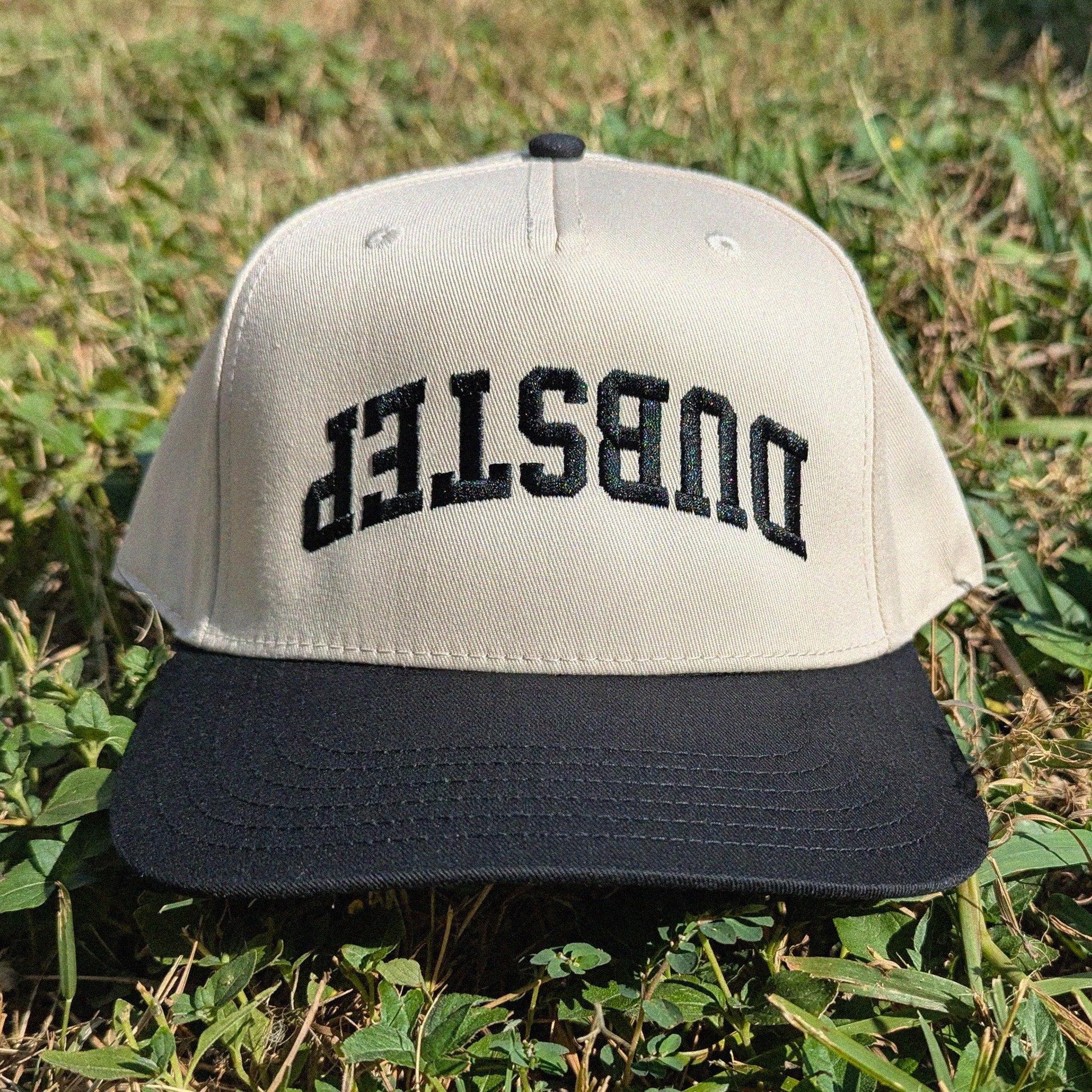 A two-tone baseball cap with a beige crown and a black curved brim, featuring the word 'DUBSTEP' embroidered in black across the front. The cap is photographed outdoors resting on green grass.