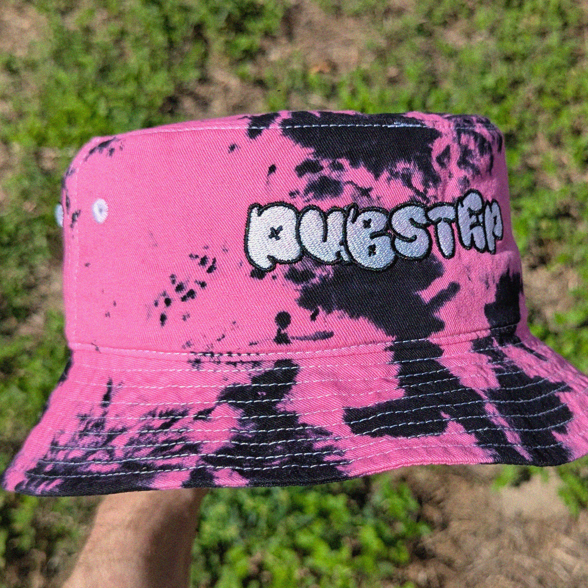 A pink and black tie-dye bucket hat with 'DUBSTEP' embroidered in a stylized white and black font on the front, held up by a hand against a blurry outdoor grassy background.