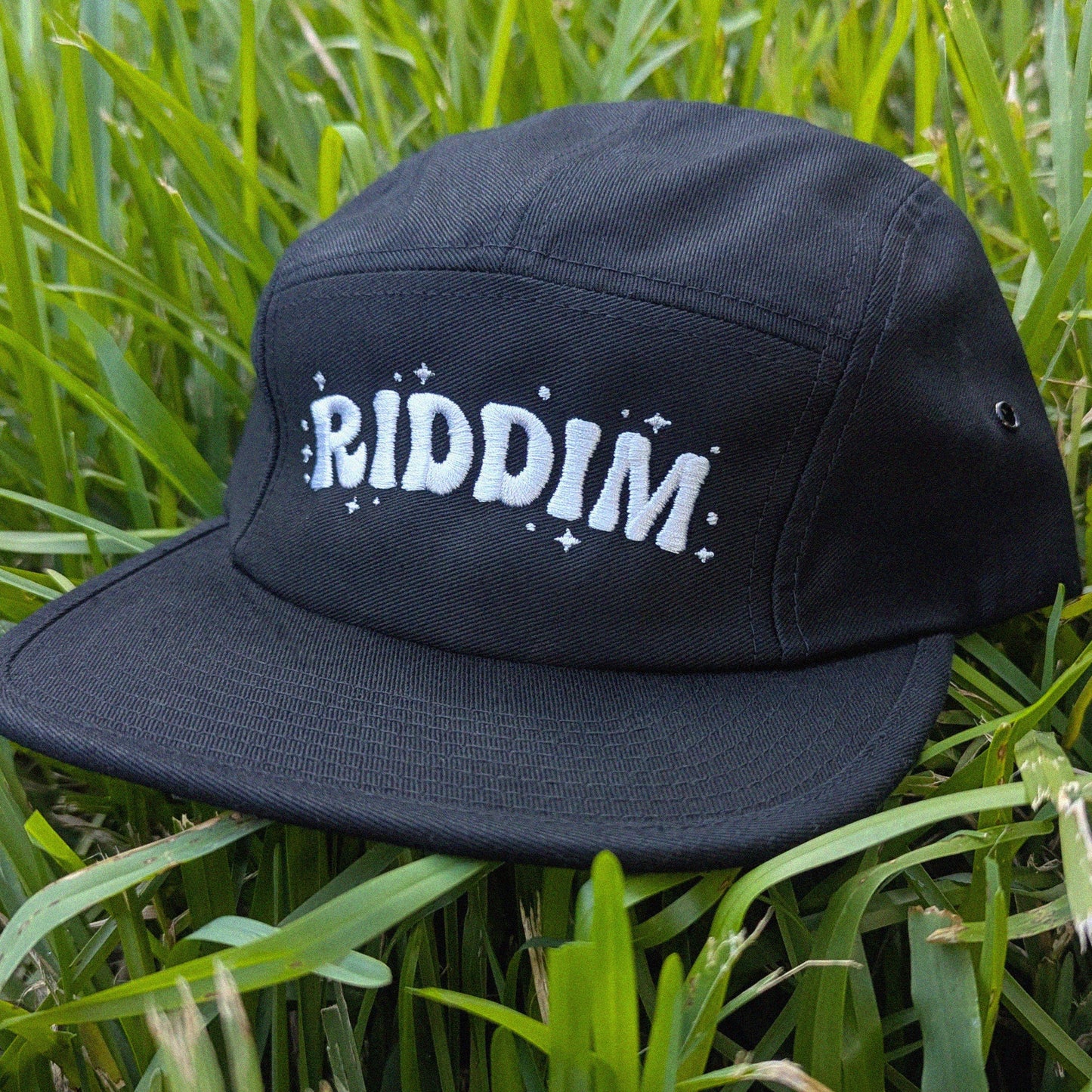 A black five-panel cap with 'RIDDIM' embroidered in white across the front, resting on green grass.