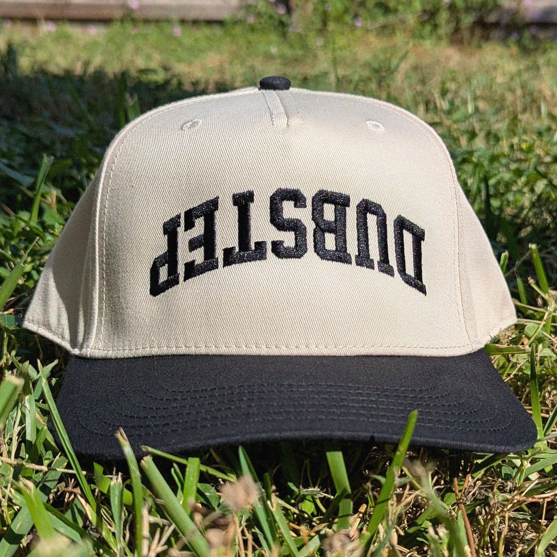 A two-tone baseball cap resting on green grass outdoors. The cap has a light beige crown with black arched embroidered text reading 'DUBSTEP' and a black flat brim.