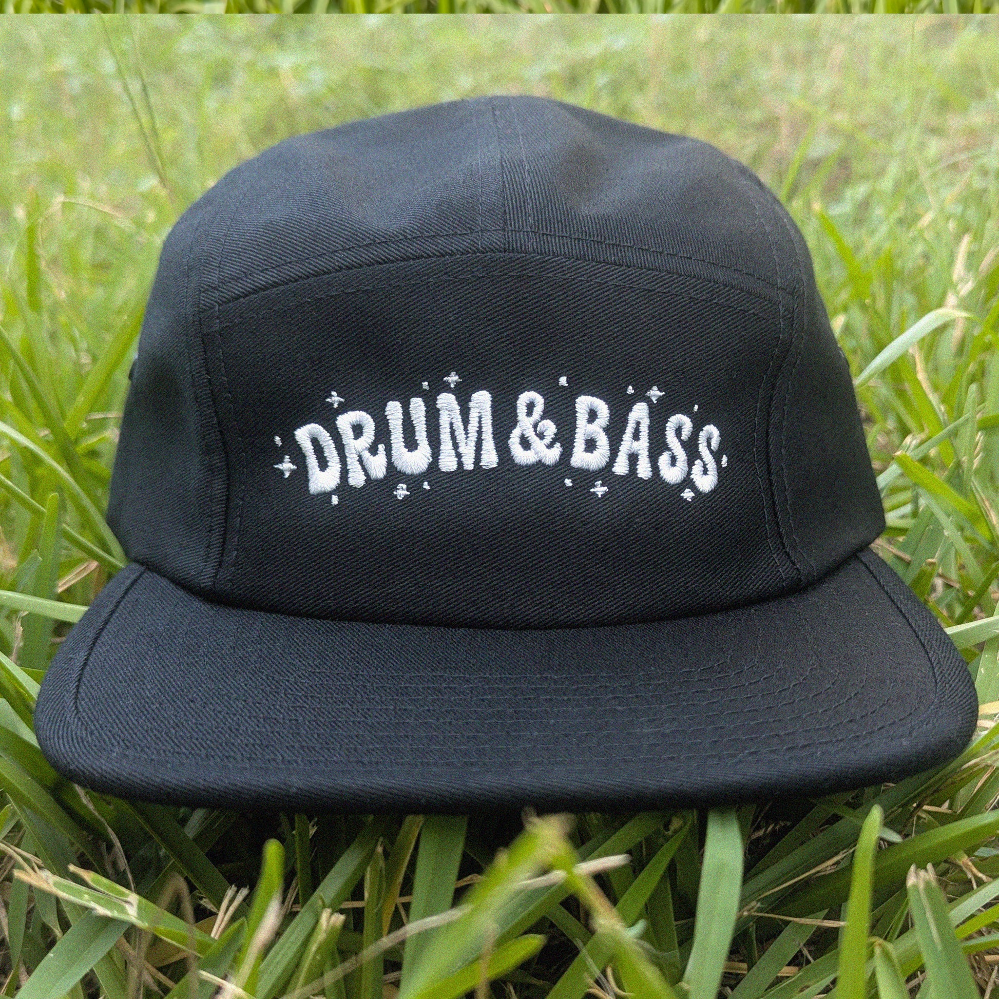 A black five-panel baseball cap with 'DRUM & BASS' embroidered in white across the front, resting on green grass.