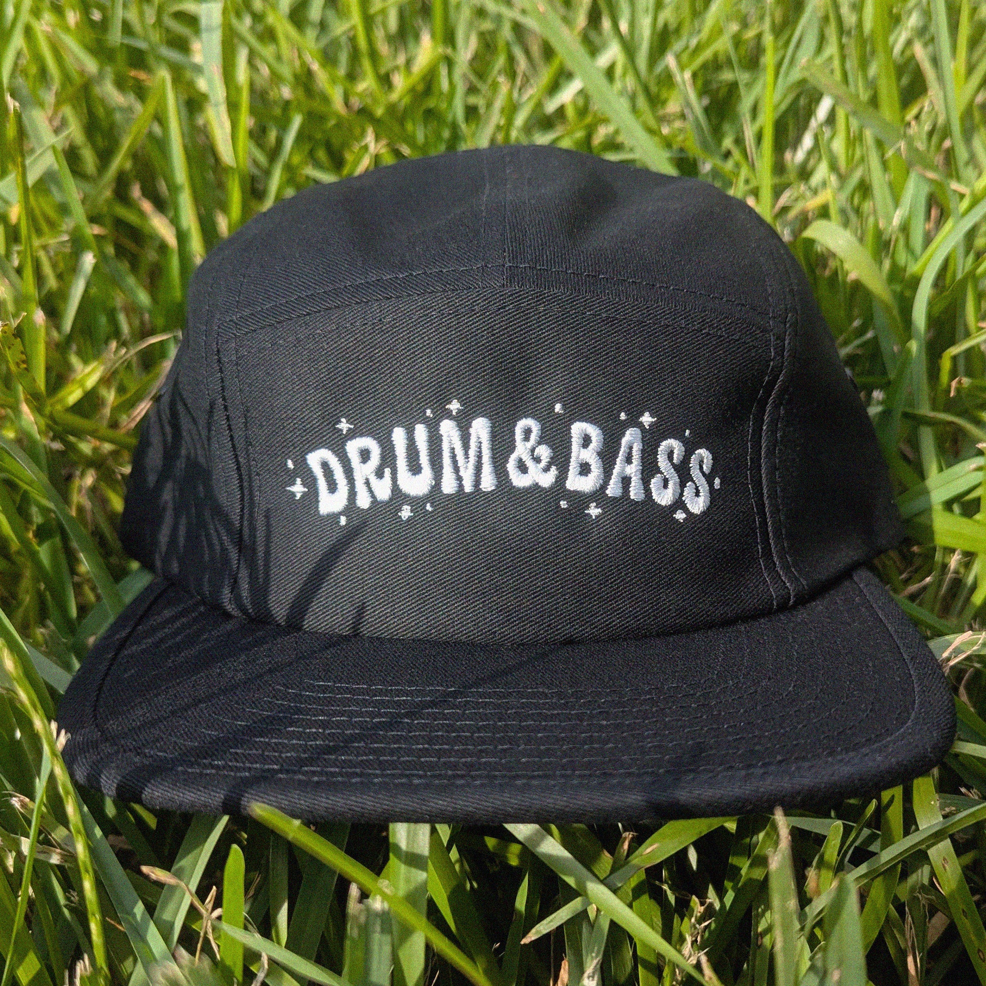 A black five-panel cap with 'DRUM & BASS' embroidered in white across the front, resting on green grass.