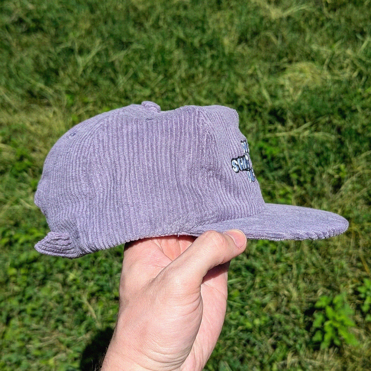 A hand holding a light purple corduroy snapback cap with a small embroidered logo on the side, set against a blurred green grass background.
