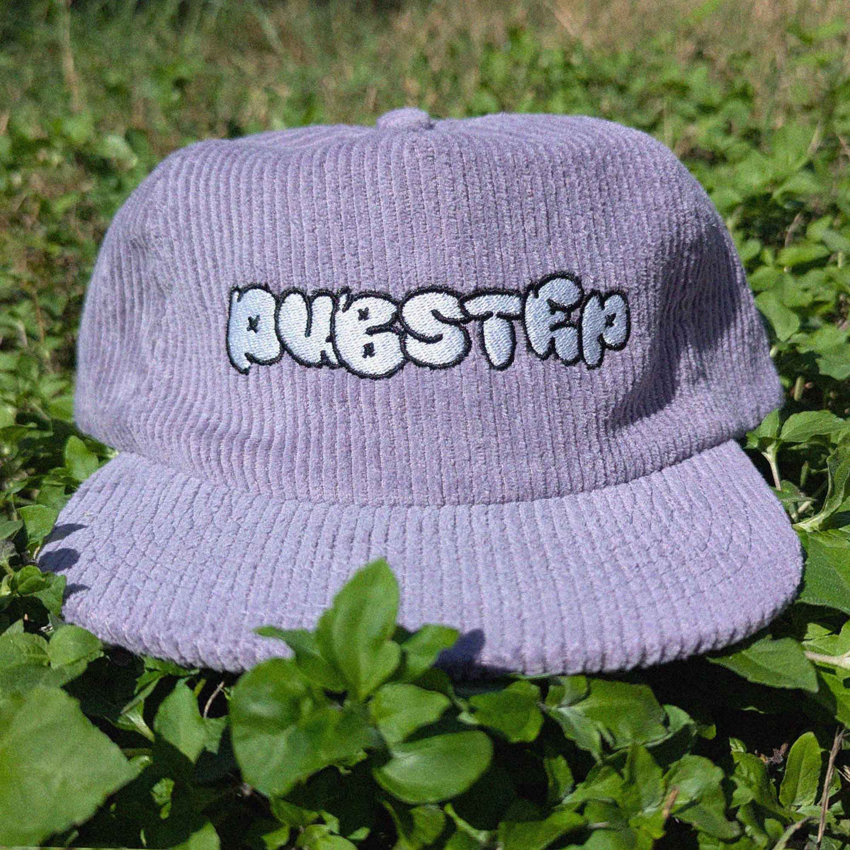 A light purple corduroy baseball cap with 'DUBSTEP' written across the front in white bubble letters with a black outline, resting on green foliage.