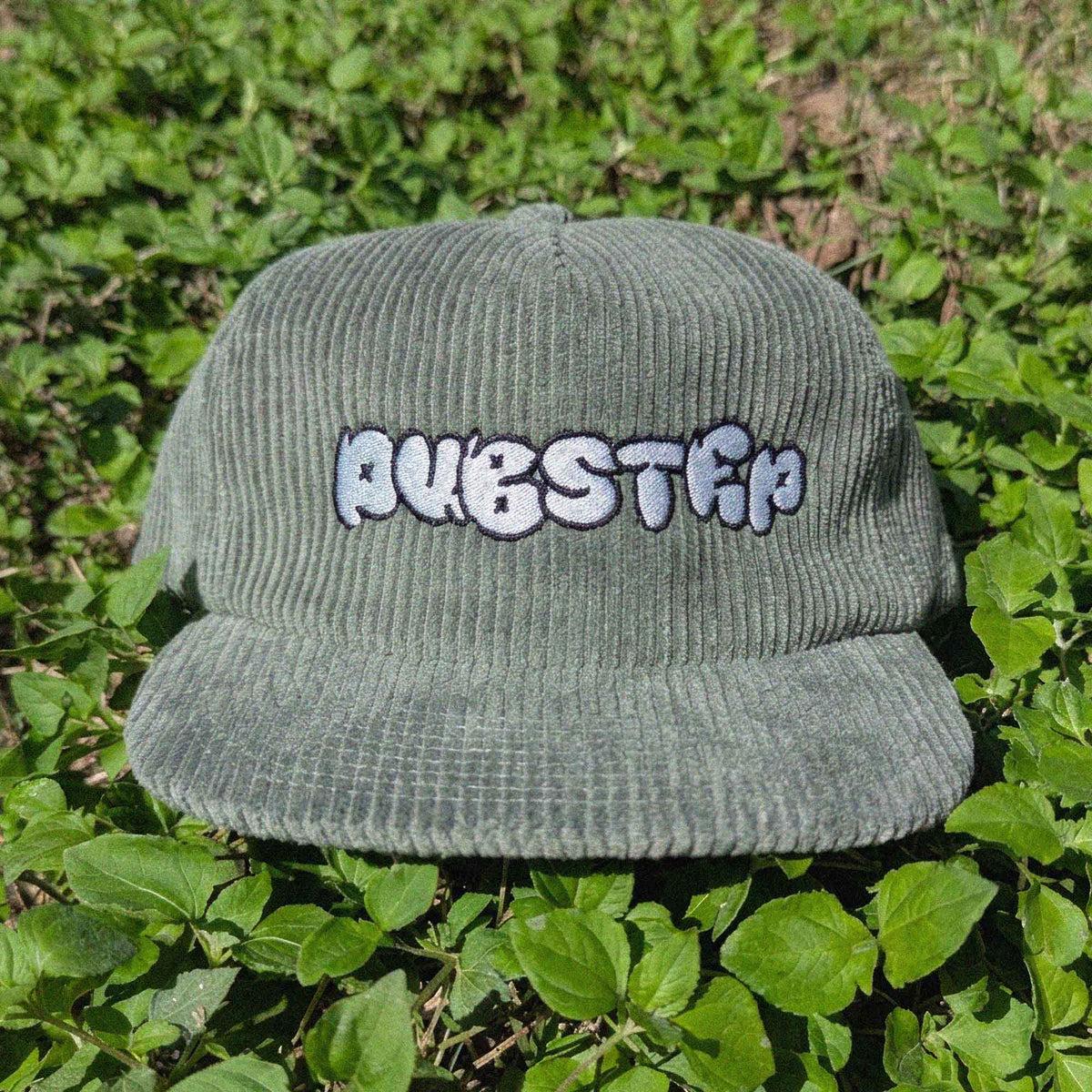 A dark green corduroy baseball cap with 'DUBSTEP' embroidered in white and black bubble letters on the front, resting on green foliage.