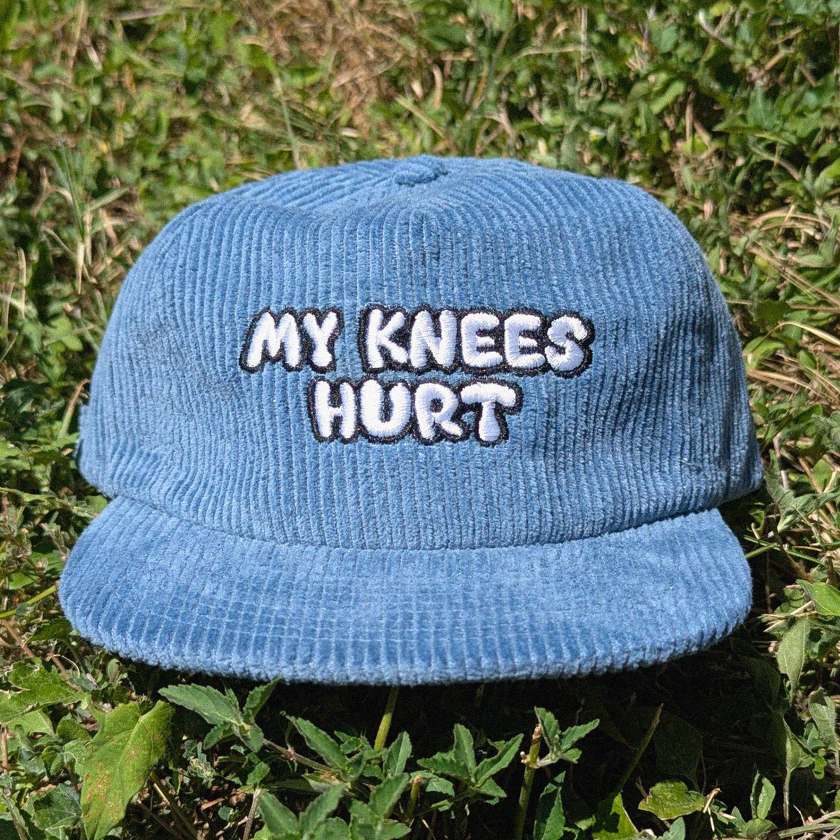 A blue corduroy baseball cap with 'MY KNEES HURT' embroidered in white and black on the front, resting on green grass.