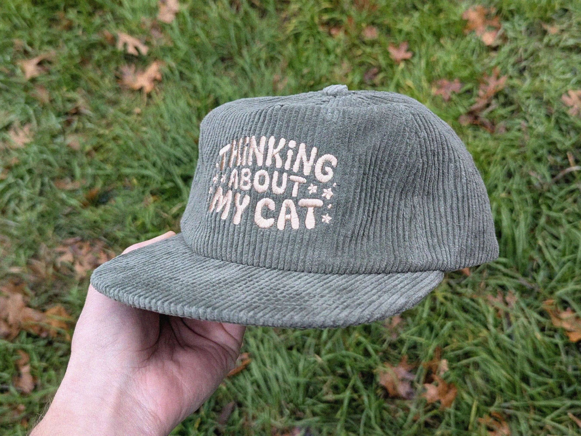 Green cap with 'Thinking About My Cat' text held in front of grass