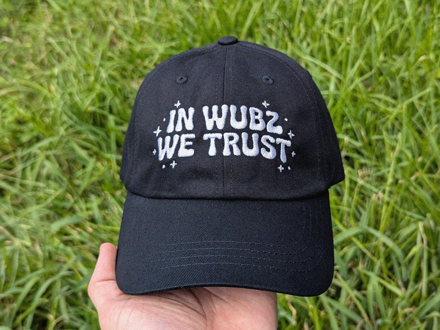 Black cap with 'IN WUBZ WE TRUST' text held in front of grass