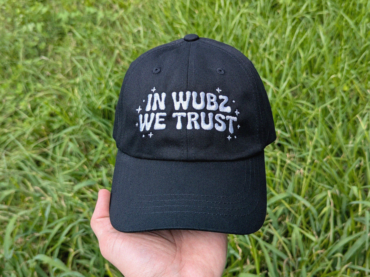 Black cap with 'IN WUBZ WE TRUST' text held in front of green grass
