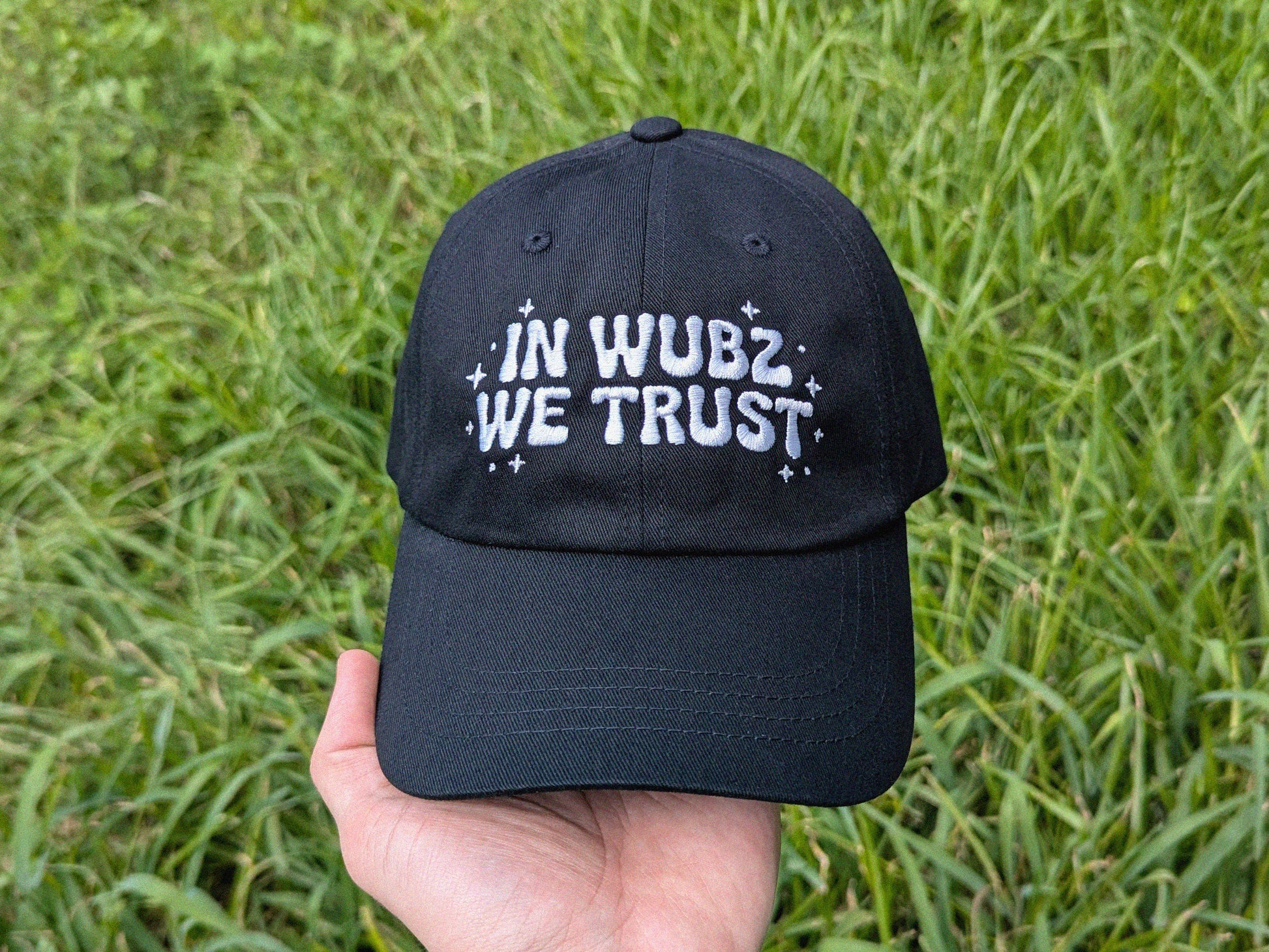 Black cap with 'IN WUBZ WE TRUST' text held in front of green grass