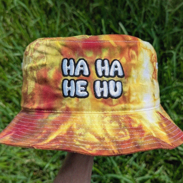 A bucket hat with a yellow, orange, and red tie-dye pattern, featuring the text 'HA HA HE HU' embroidered on the front. The hat is held up by a hand against a background of green grass.