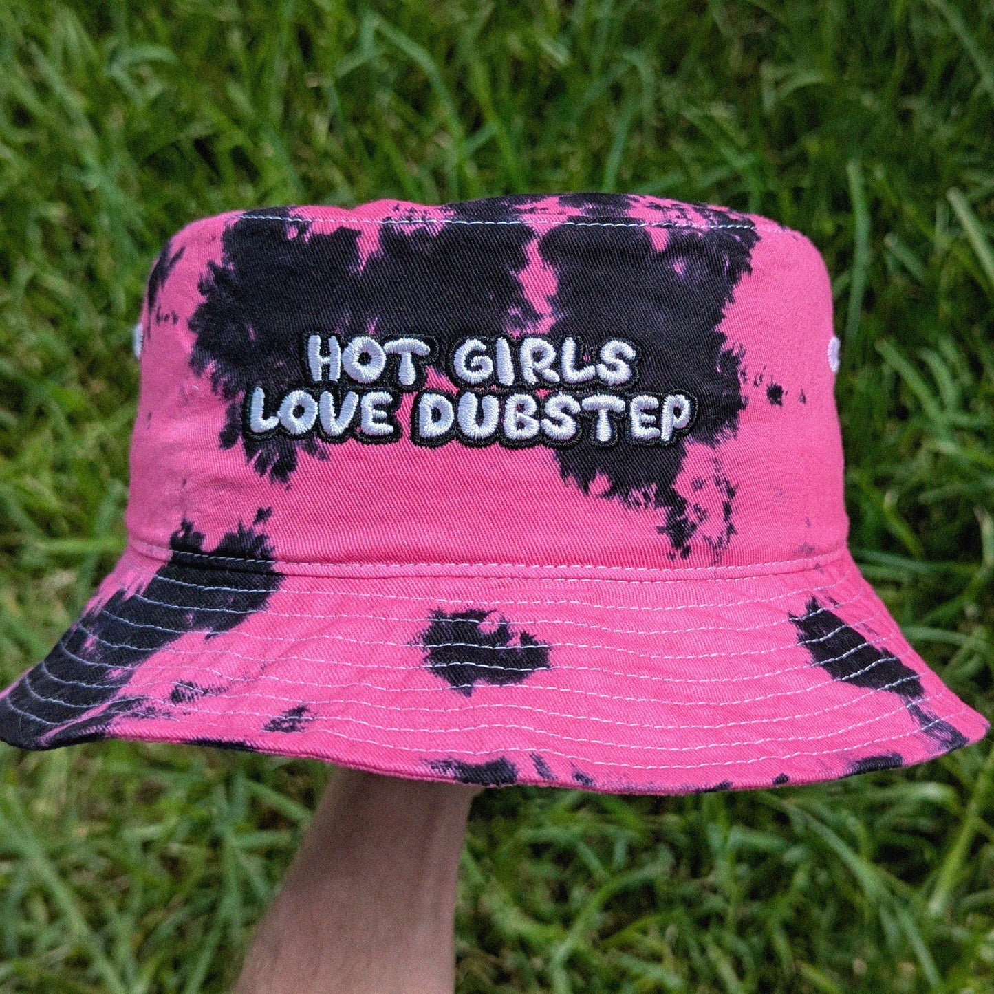 A pink and black tie-dye bucket hat with the text 'HOT GIRLS LOVE DUBSTEP' embroidered on the front, held up by a hand against a background of green grass.