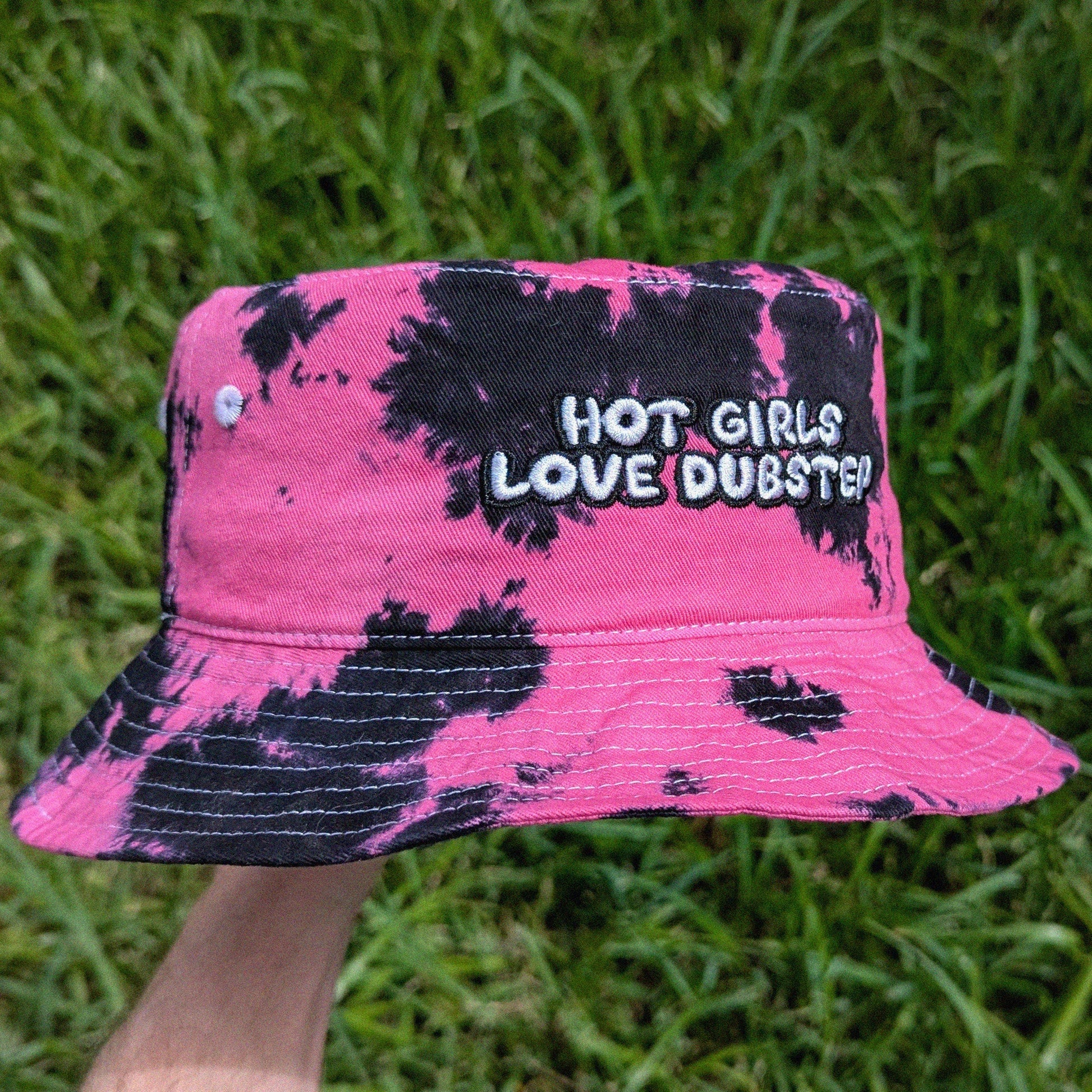A pink and black tie-dye bucket hat with the text 'HOT GIRLS LOVE DUBSTEP' embroidered on the front, held up by a hand against a background of green grass.