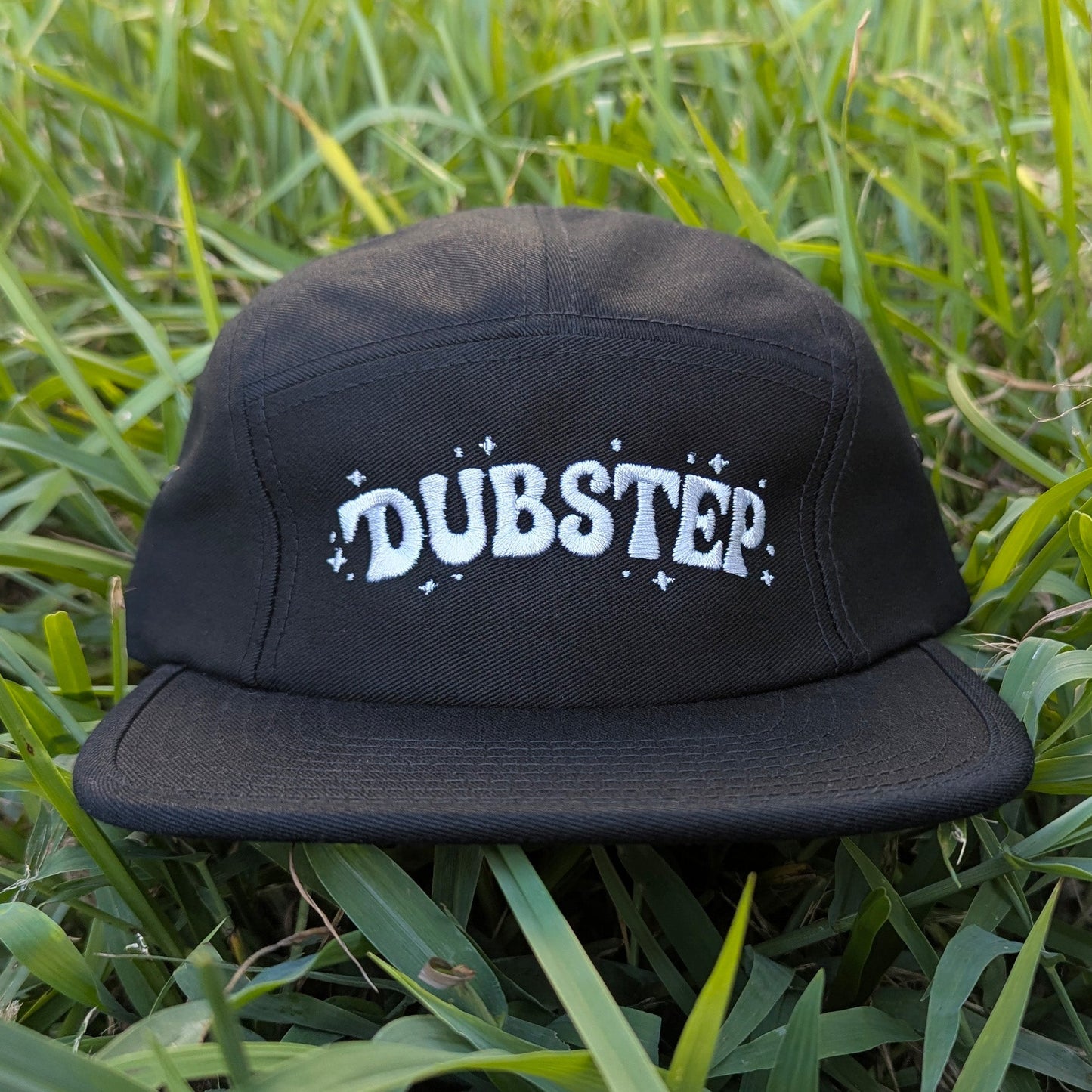 A black flat-brim cap with 'DUBSTEP' embroidered in white across the front, resting on green grass.