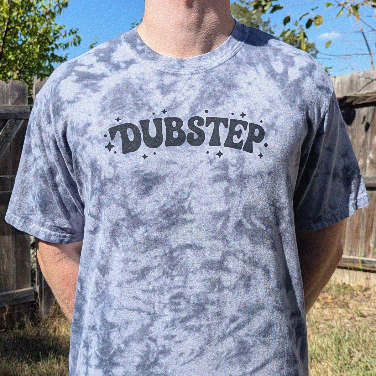 A person wearing a gray tie-dye t-shirt with the word 'DUBSTEP' printed across the chest in a dark gray stylized font, set against an outdoor background with a wooden fence and trees.