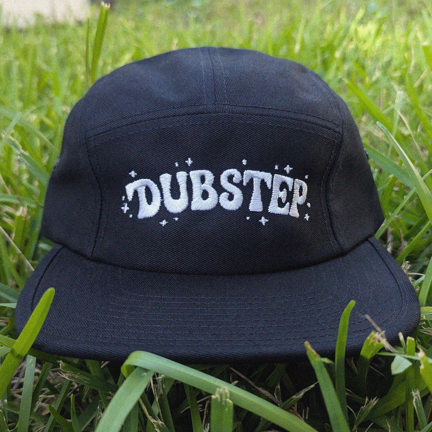 A black five-panel cap with 'DUBSTEP' embroidered in white across the front, resting on green grass.