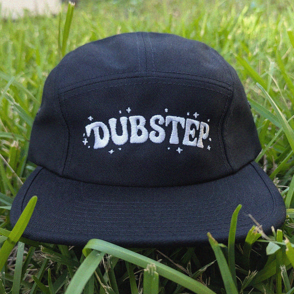 A black five-panel cap with 'DUBSTEP' embroidered in white across the front, resting on green grass.