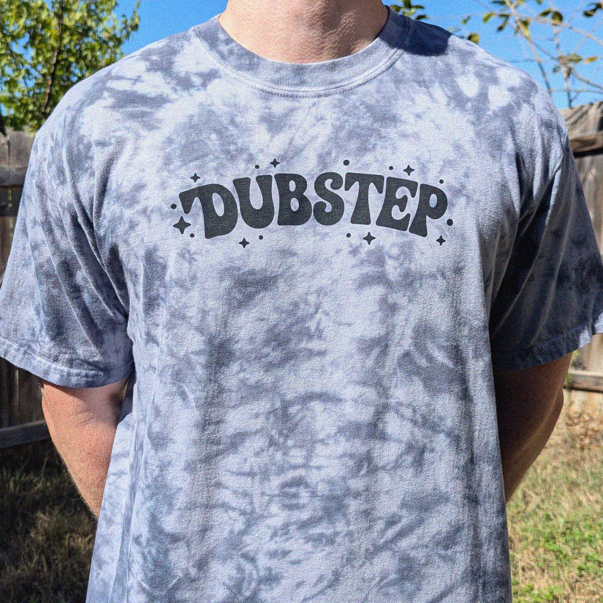 A person wearing a gray tie-dye t-shirt with the word 'DUBSTEP' printed in black across the chest, photographed outdoors in a backyard setting.