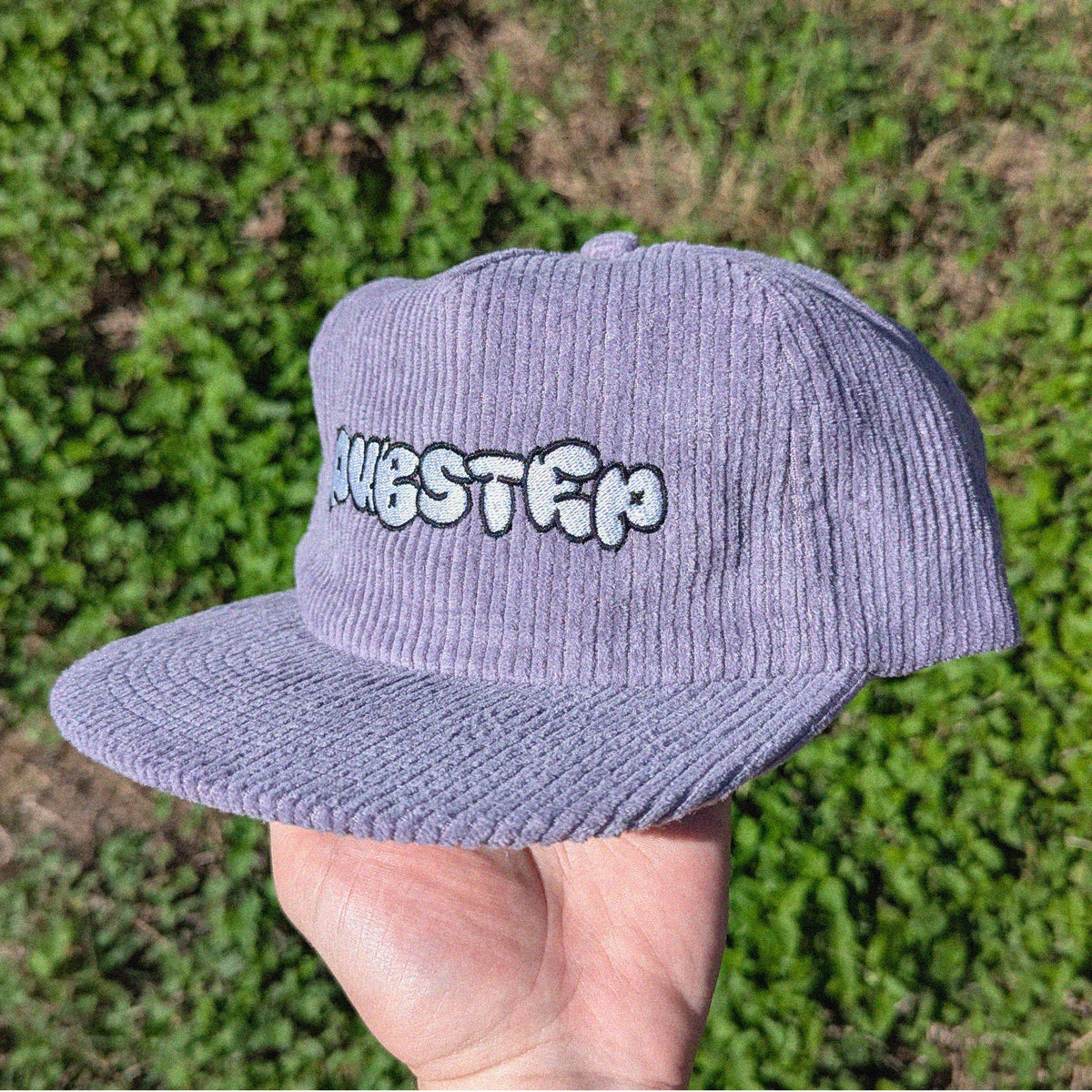 A lavender corduroy snapback cap with 'DUBSTEP' embroidered on the front, held in a hand against a blurred green grass background.