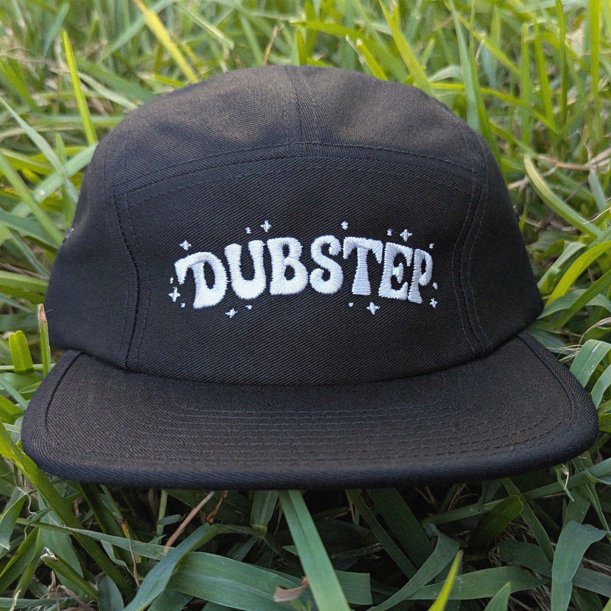 A black five-panel cap with 'DUBSTEP' embroidered in white across the front, resting on green grass.