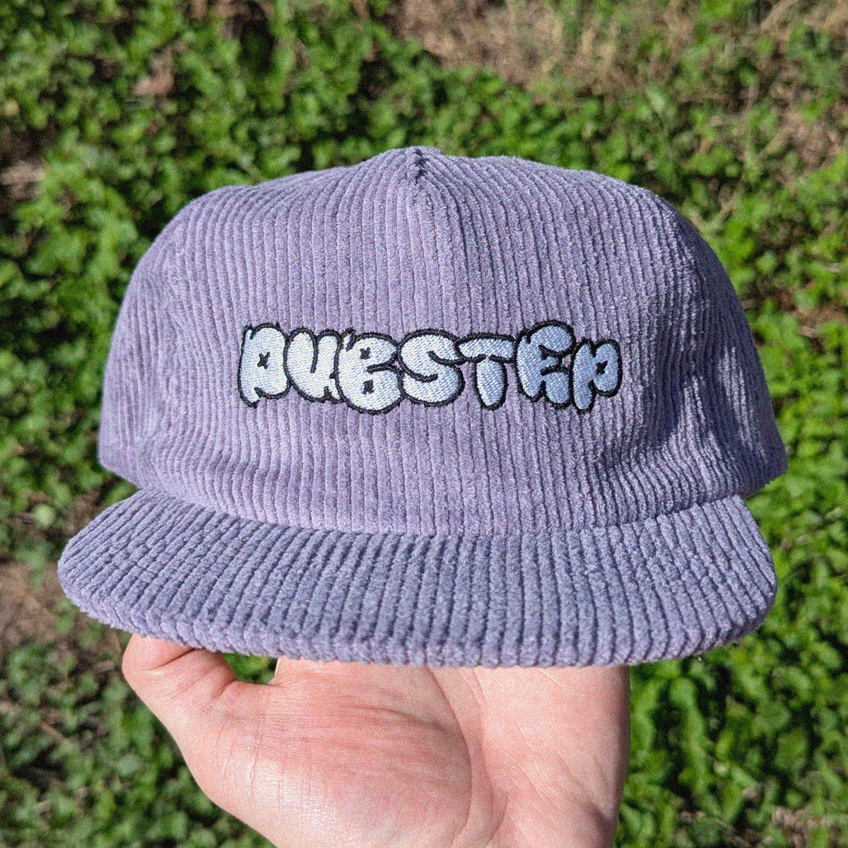 A lavender corduroy baseball cap with 'DUBSTEP' embroidered in white and black bubble letters on the front, held in a hand against a blurred green grass background.