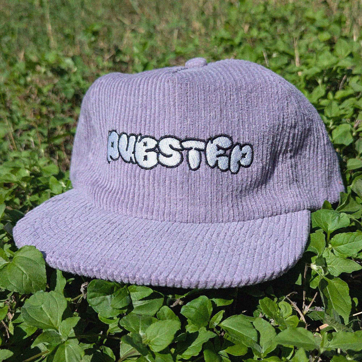 A light purple corduroy baseball cap with 'DUBSTEP' embroidered in white and black bubble letters on the front, resting on green foliage.