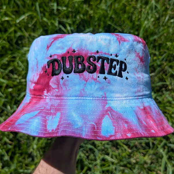 A bucket hat with a pink and blue tie-dye pattern, featuring the word 'DUBSTEP' embroidered in black across the front, held up by a hand against a background of green grass.