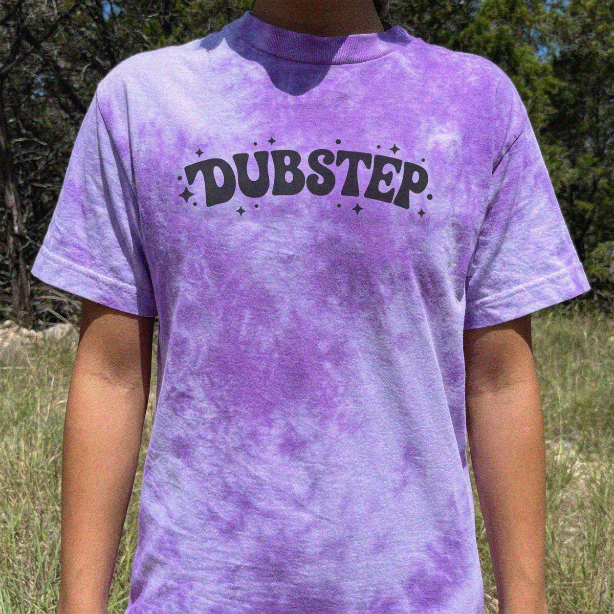 A person wearing a purple and lavender tie-dye t-shirt with the word 'DUBSTEP' printed in black across the chest, standing outdoors in a grassy field with trees in the background.
