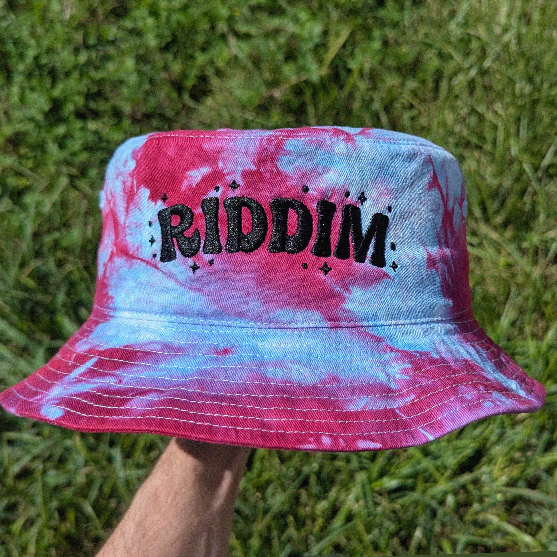 A bucket hat with a pink and blue tie-dye pattern, featuring the word 'RIDDIM' embroidered in black across the front. The hat is held up by a person's hand against a background of green grass.