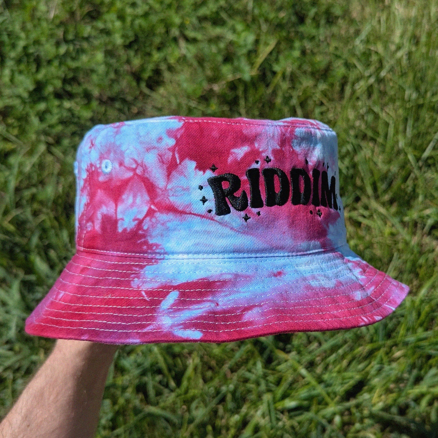 A hand holding a red, white, and blue tie-dye bucket hat with the word 'RIDDIM' printed in black across the front, set against a background of green grass.