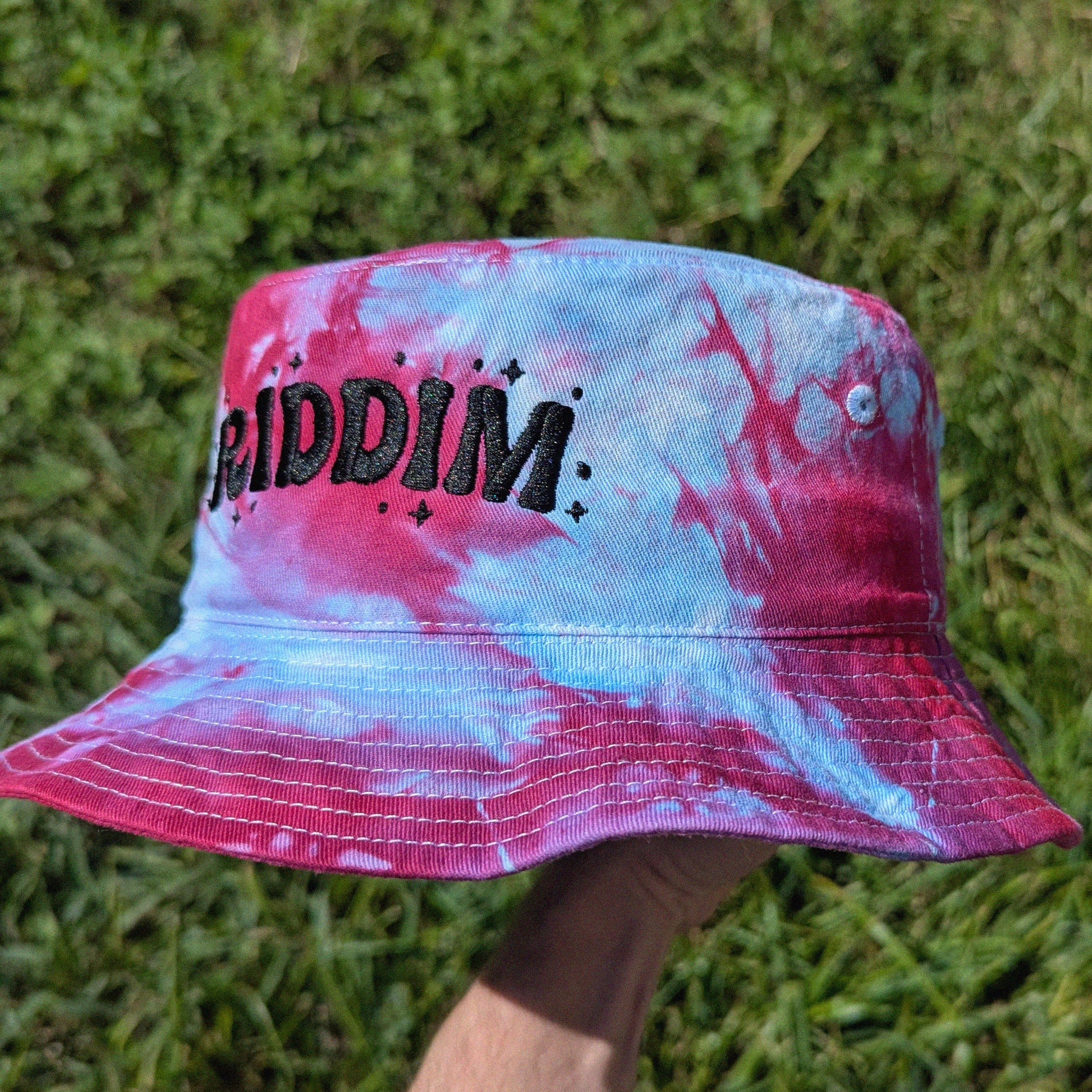 A bucket hat with a pink, blue, and white tie-dye pattern, featuring the word 'RIDDIM' in black glittery letters on the front. The hat is being held by a hand against a background of green grass.