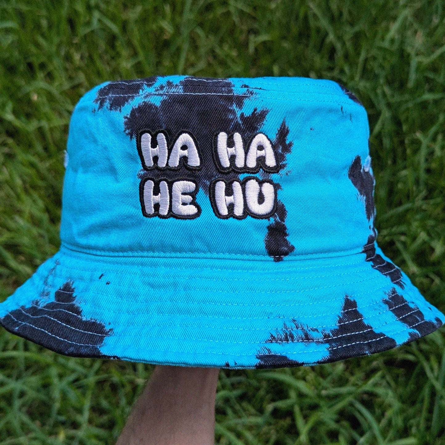 A black and teal tie dye bucket hat with embroidered text reading 'HA HA HE HU' on the front, held up outdoors against a background of green grass.