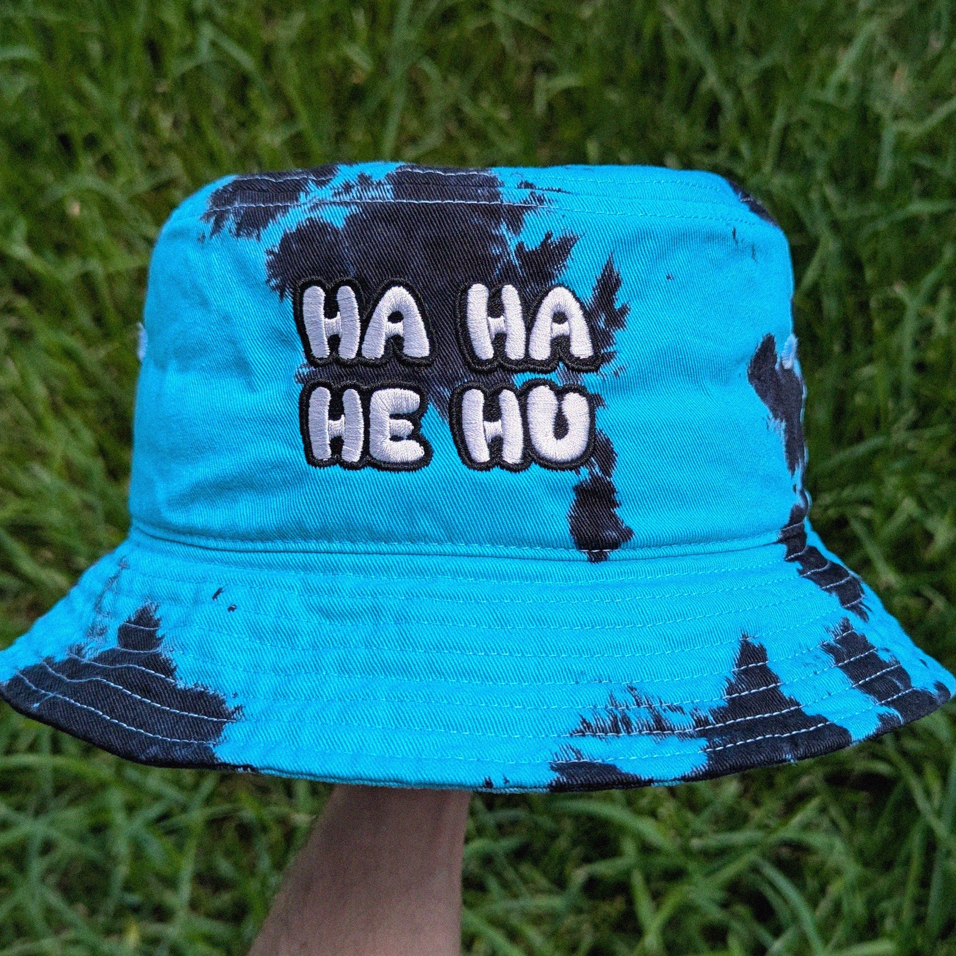 A black and teal tie dye bucket hat with embroidered text reading 'HA HA HE HU' on the front, held up outdoors against a background of green grass.