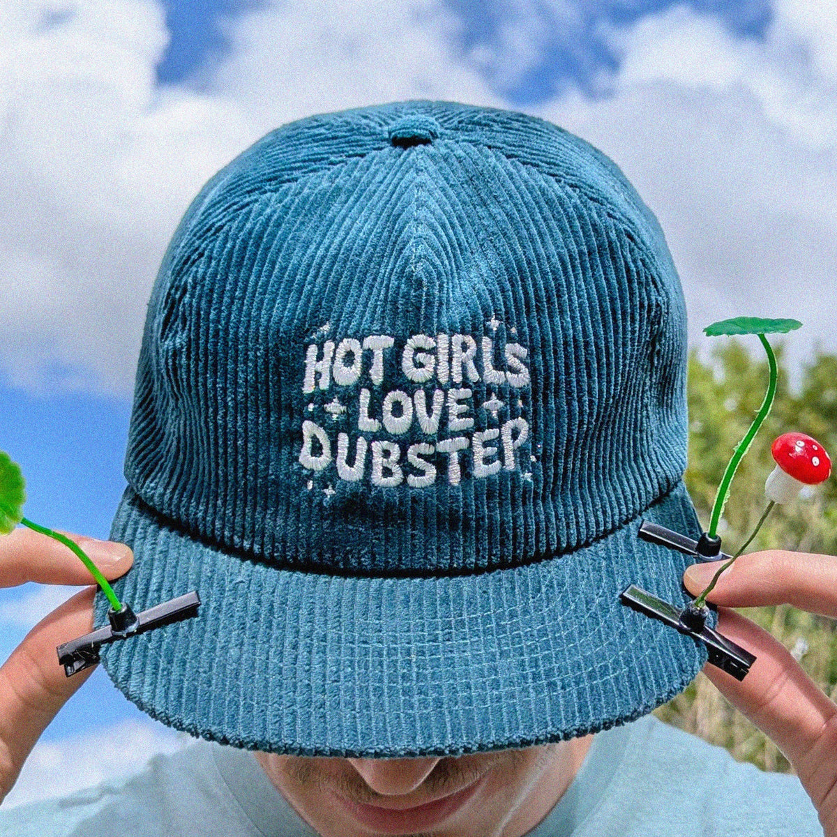 A person outdoors under a blue sky wearing a blue corduroy baseball cap with the phrase 'HOT GIRLS LOVE DUBSTEP' in white embroidered text.